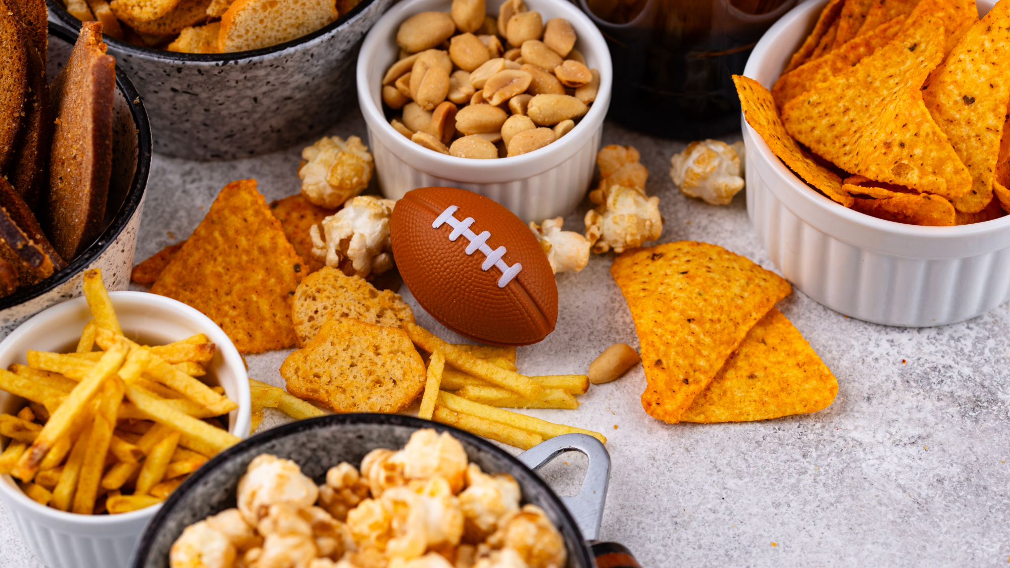 Game day snack spread with tortilla chips, popcorn, peanuts, fries, and a football centerpiece, styled for a Super Bowl watch party upsell.